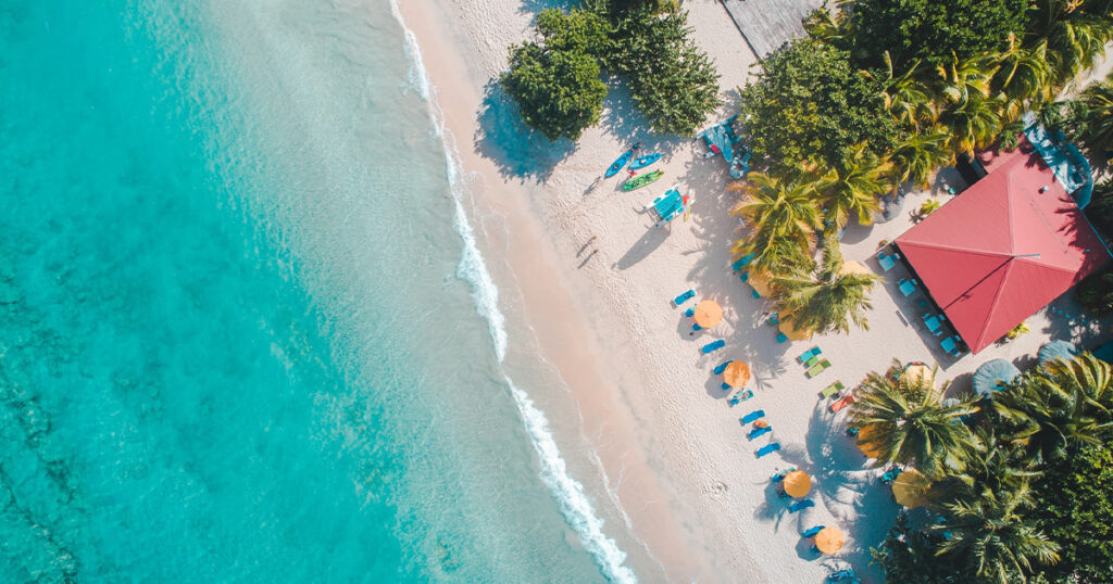 team health starts on vacation. A birds eye view image of a beach with lounge chairs and palm trees. A red roofed building sits in the trees