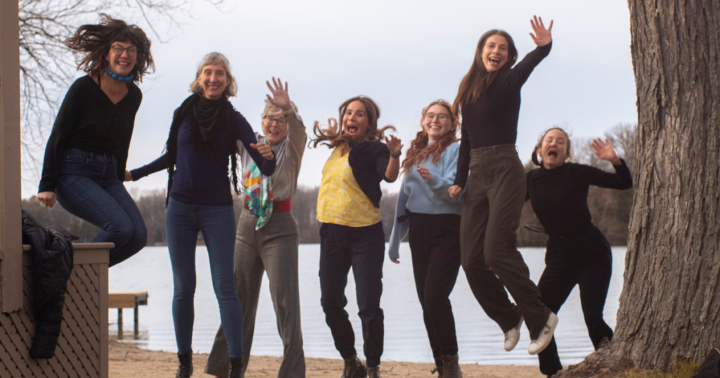 7 strong women from Neolé jumping in the air in front of a lake, although only 2 of them are in the air. Wee!