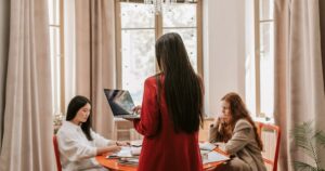 a women standing with her back facing us holding a laptop with 2 people sitting on both sides of her, at a table in front of her. Focusing on work-life balance