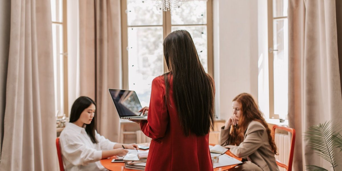 a women standing with her back facing us holding a laptop with 2 people sitting on both sides of her, at a table in front of her. Focusing on work-life balance